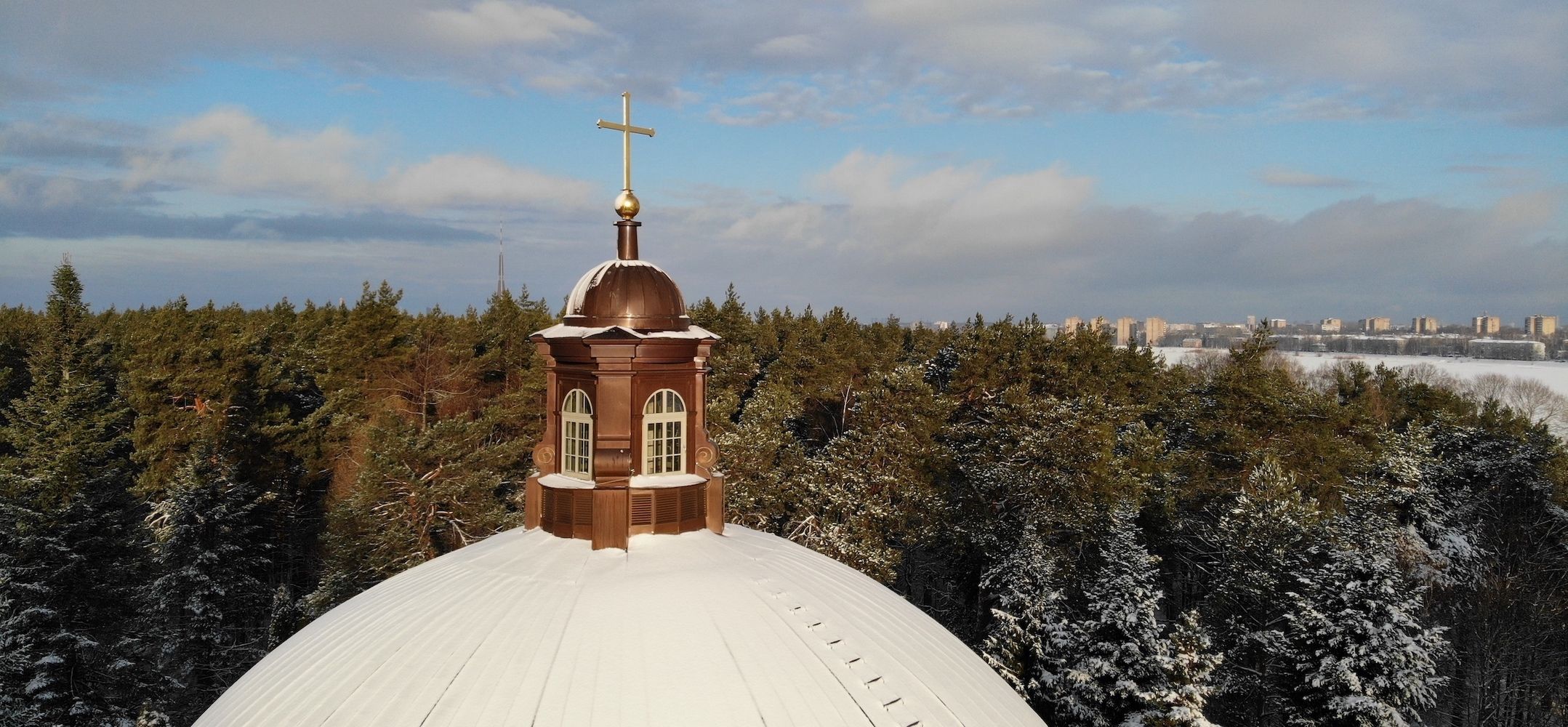 Snow-covered dome and steeple with a golden cross rising above a winter forest.